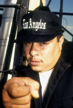 A man with a faint mustache wearing a Los Angeles baseball cap points at the camera close-up.