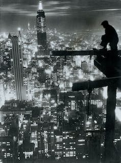 Man sits on construction scaffolding overlooking New York City skyline.