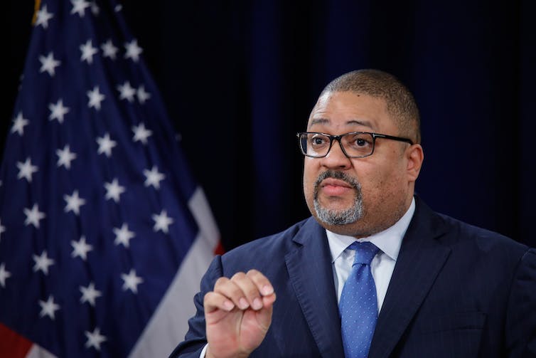 A black man dressed in a business suit gestures with his hand as he stands near an American flag.