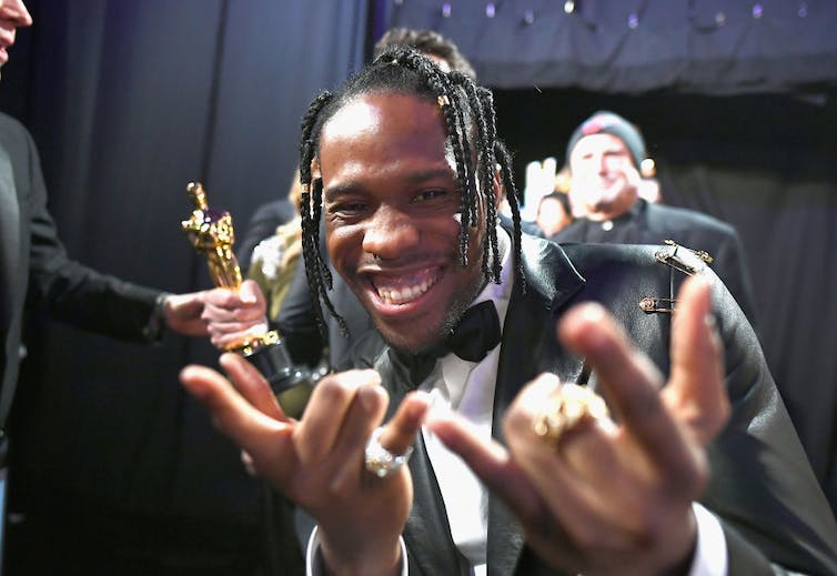 Smiling man with dreadlocks poses with 'shocker' hand gestures.