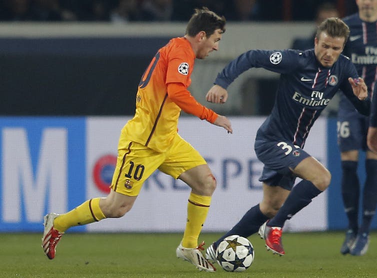 A man in a yellow and orange soccer kit dribbles a ball past a player in blue.