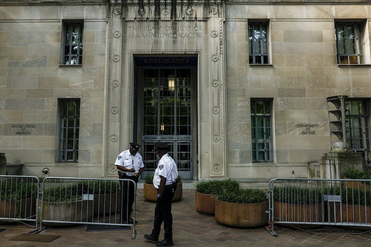 Two men in police uniforms stand outside a limestone building fronted by police barricades.