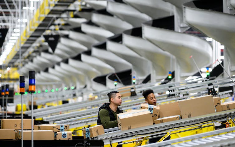 Two young men stand beside a conveyor belt with boxes on it. Several more belts just like it are behind them.