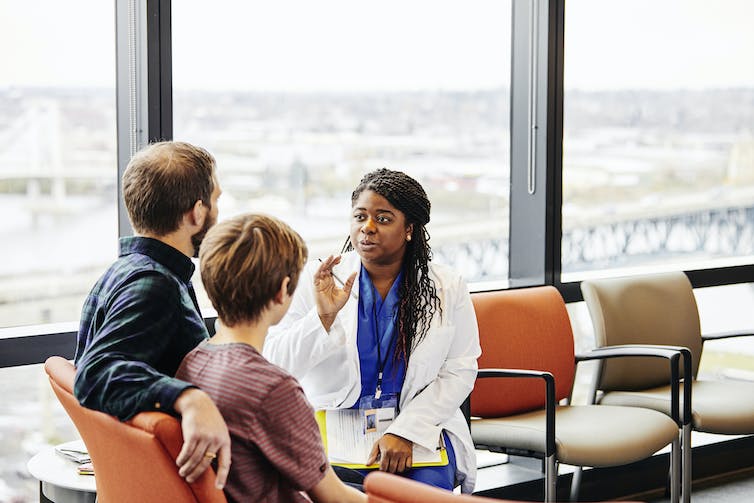 A woman in blue scrubs and a white doctor's coat chats with a man and boy in a room with views out over a city.