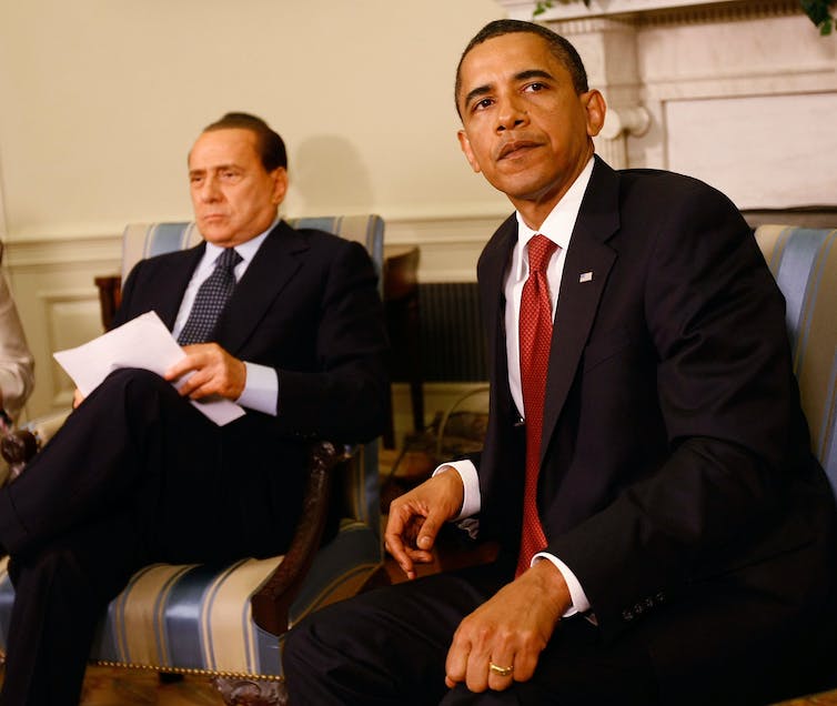 A glum looking man looks off to the side next to a similarly downcast man shuffling papers.