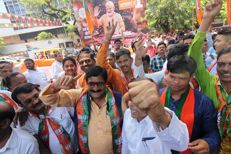 A crowd of men smile, raise their hands and wear orange, in front of a political poster of an older man with a white beard and hair.