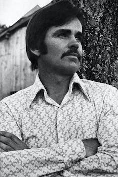 Black and white photo of man with mustache folding his arms.