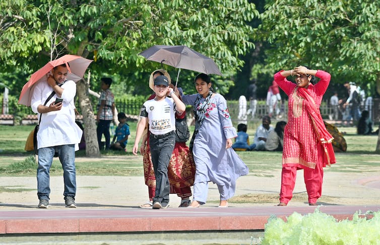 Three adults walk under umbrellas sheltering them from the sun. A woman without an umbrella shades her eyes with her hands on a hot day, and a boy wears a cap.