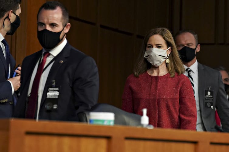 A woman in a dress walks behind a wooden podium. She is flanked by two men in suits and all three people are wearing masks.