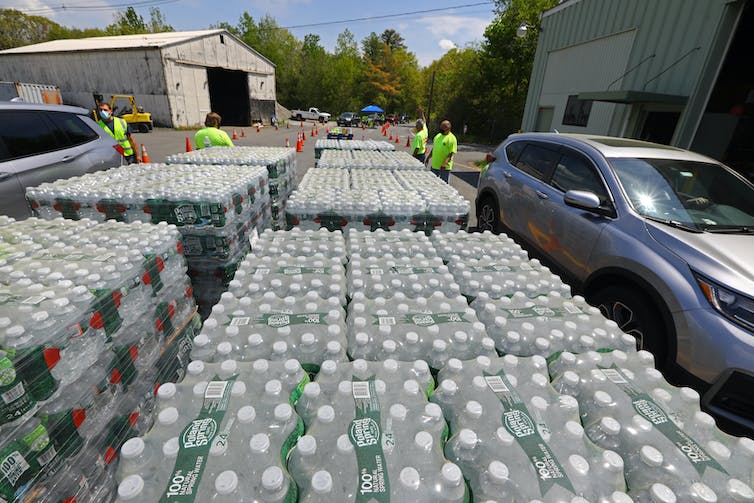 Several pallets of bottled water sit while people prepare to put it into the trunks of SUVs picking it up.