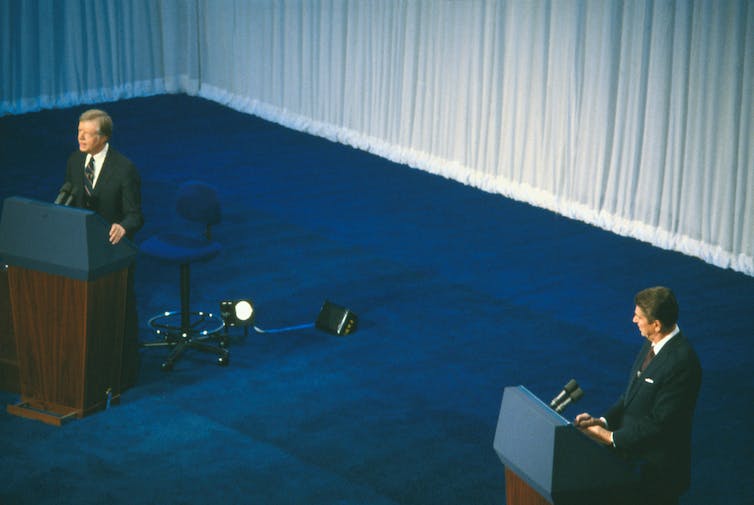 Two suited men stand behind individual lecterns. The man on the left speaks as the man on the right looks in his direction.