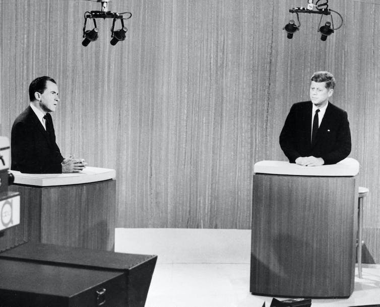 Two suited men stand behind individual lecterns, resting their hands atop the stand.