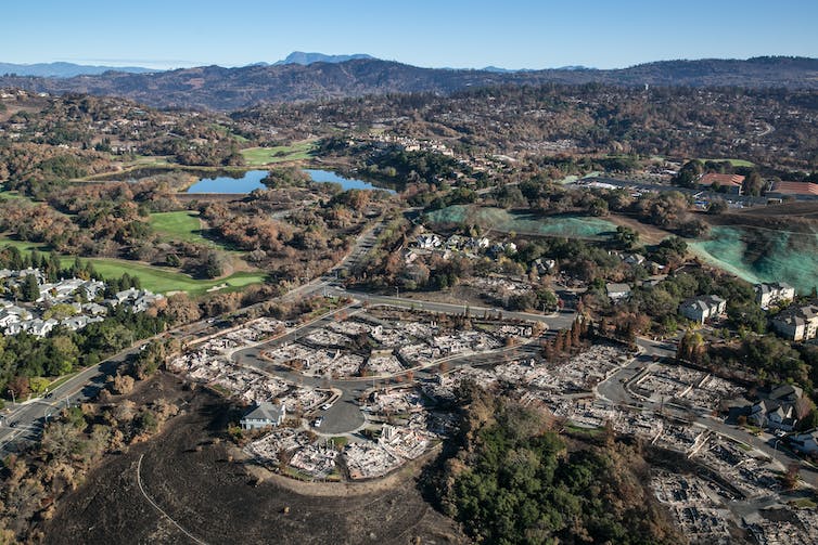 A landscape view across a neighborhood with gold courses, lakes and hills in the background. In the foreground is burned cul de sac that appears to be at the edge of the city.