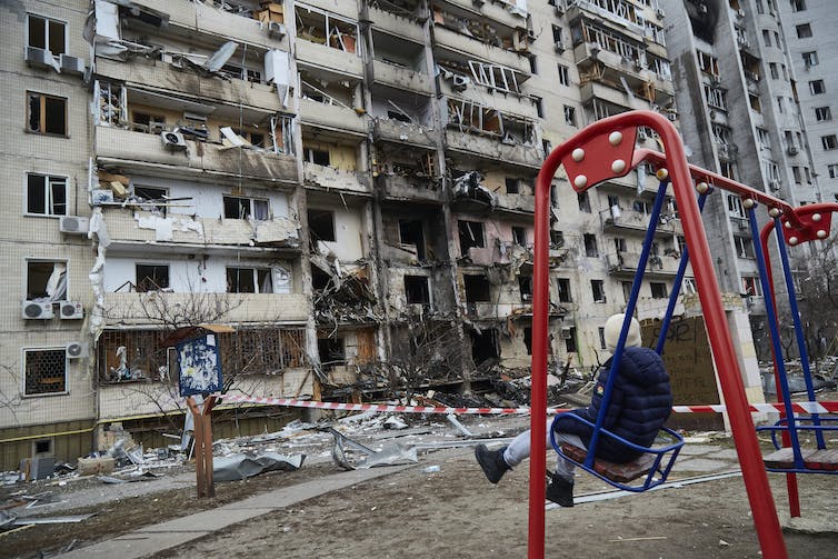 A child swings in front of a destroyed, burned looking building.