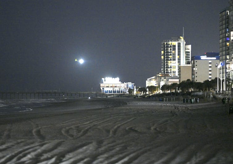 An illuminated spy balloon flies in the night sky near lit buildings and above a sanded section of a beach near the ocean.