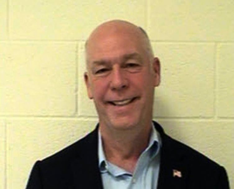 A balding man in a dark blazer and blue shirt standing against a yellow wall.