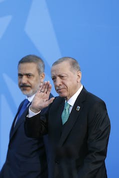 A man in a dark suit waves in front of a blue screen.