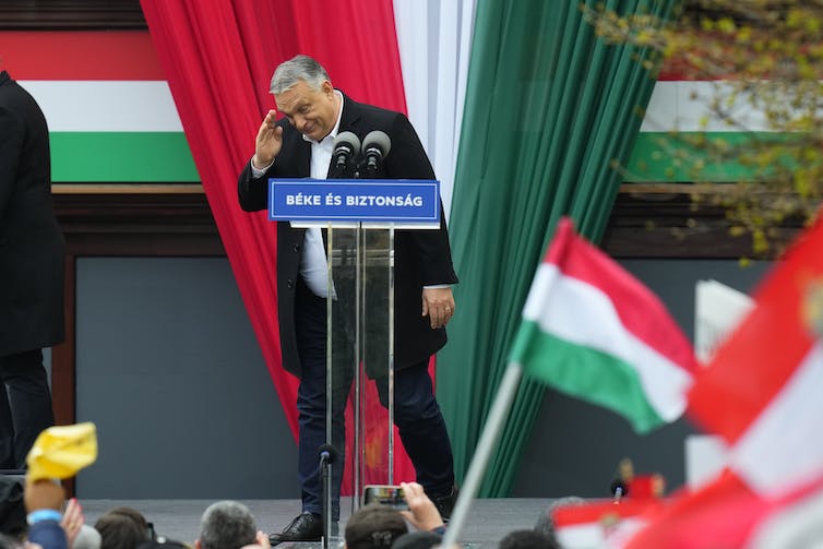 Viktor Orban salutes to a crowd of people while he stands on a podium, with red, white and green flags around him.