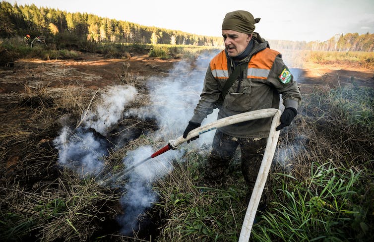 A volunteer with no mask or protective gear holds a fire hose as he fights an underground fire in a peat bog. The open bog is behind him, rimmed by forest.