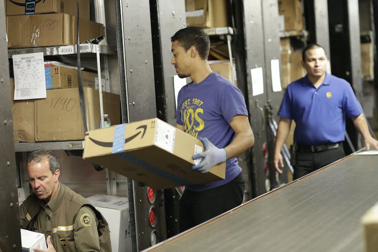 A worker carries an Amazon box as another checks over a box and address.