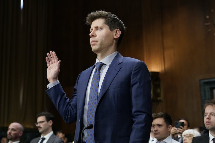 A man in a business suit stands with his right hand raised in a wood-paneled room.