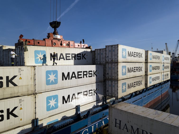 A person runs across the top of a Maersk shipping container on a smaller container ship while more containers are loaded.