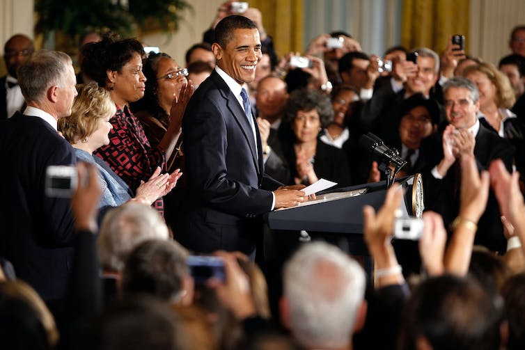 A smiling man speaks while standing at a lectern in a room full of smiling people.