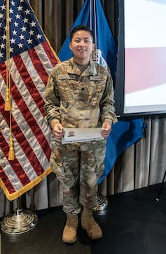 A woman dressed in a military uniform stands in front of an American flag.