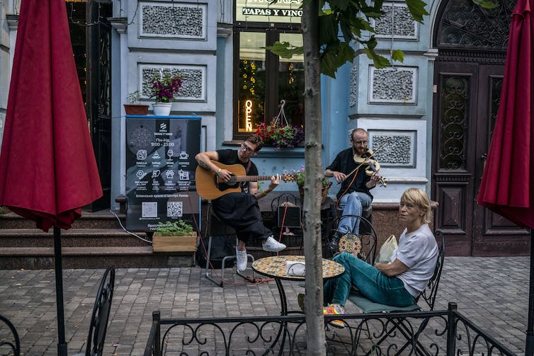 Two men play a guitar and a violin on an empty looking street, with a woman sitting in a chair nearby.