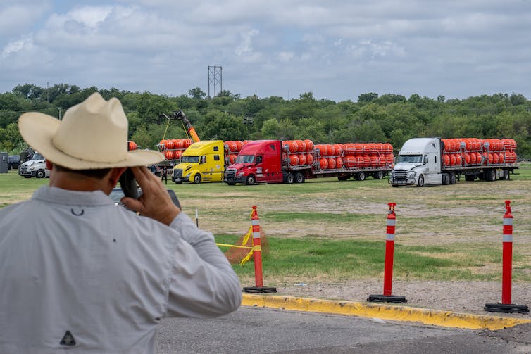 A person with a cowboy hat is seen from behind, talking on the phone as he looks at large trucks with big orange buoys lined up behind them.