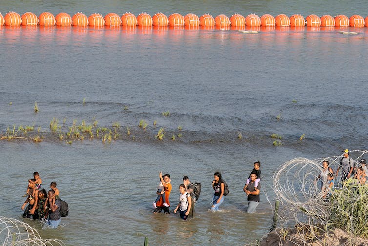 A group of people are seen walking through knee-deep water, in front of a long row of large orange circular buoys.