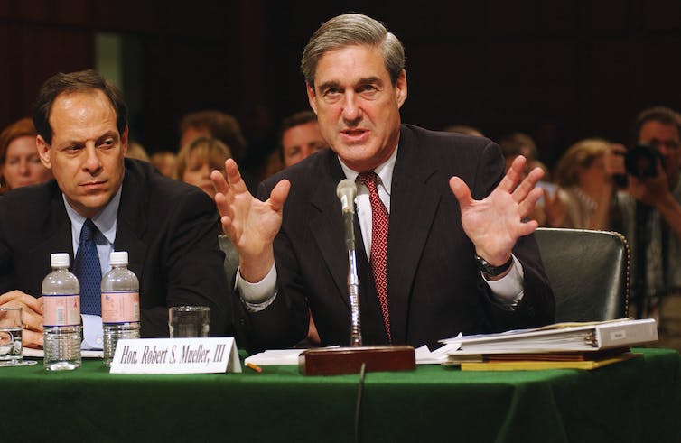 Two men in suits sitting at a table, with one talking and gesturing with his hands.