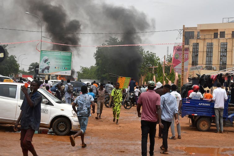 Billowing black smoke emerges from a building behind a crowd of mostly men.