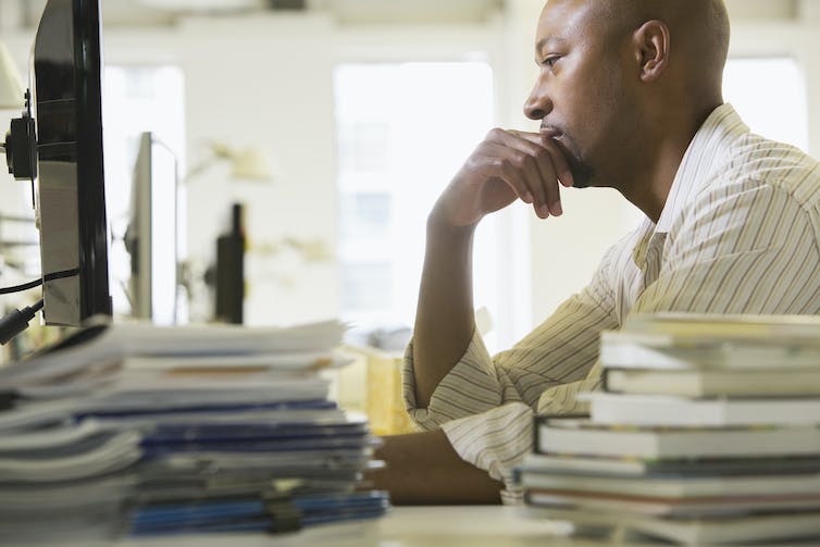 A man at a computer, chin resting on hand, with publications on the desk around him.