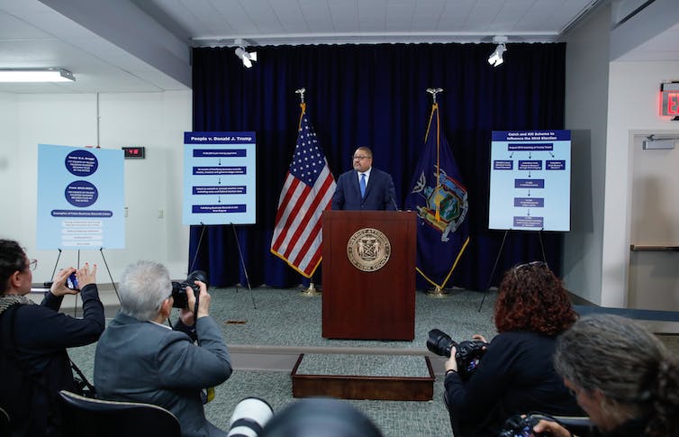 A man in a suit talks at a lectern in the front of a meeting room, with an American flag behind him.