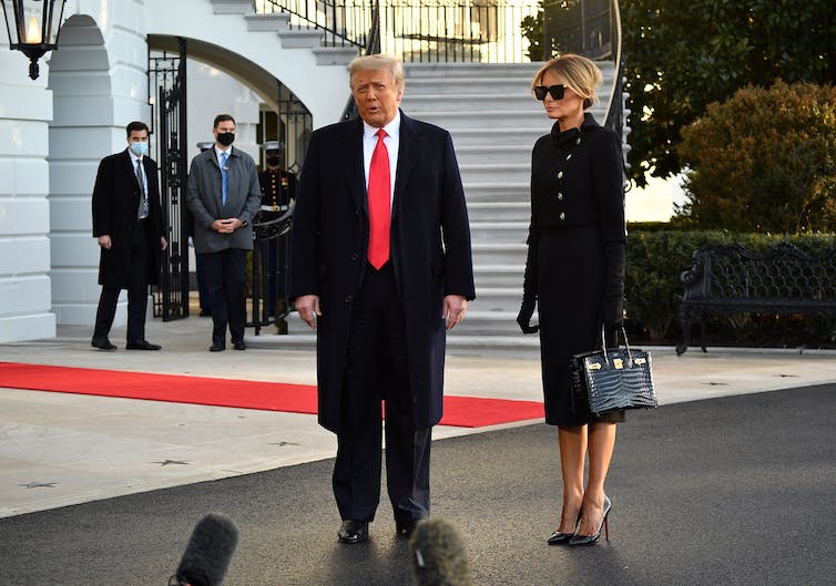 A man in a dark coat and red tie stands next to a woman in dark coat and sunglasses, talking.