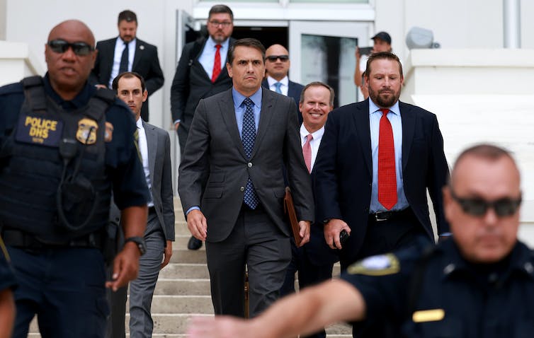 A group of men in suits walk down steps, flanked by people in black police uniforms.