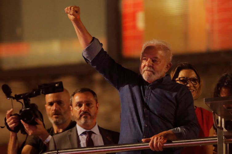 A man standing, gripping a metal rail with one hand, the other hand raised with a fist, in the nighttime.