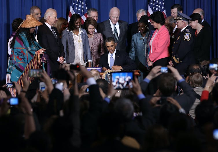 A man wearing a suit and tie sits while he signs a document. Suited men stand around him watch as he signs.