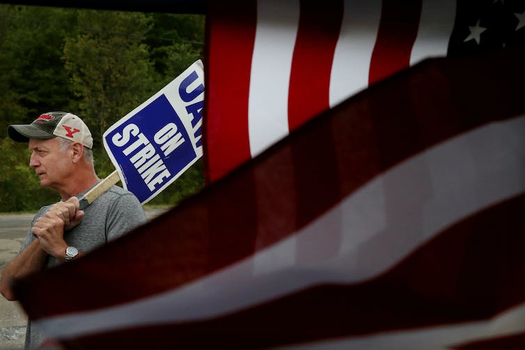 Man carries a 'UAW on strike' picket sign, enveloped in an American flag.