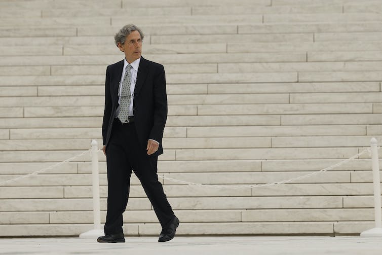 A white man dressed in a dark business suit is walking on marbled stairs.