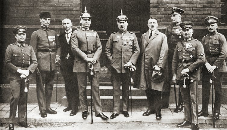 Nine men, most dressed in old-fashioned military uniforms. standing together outside a building.