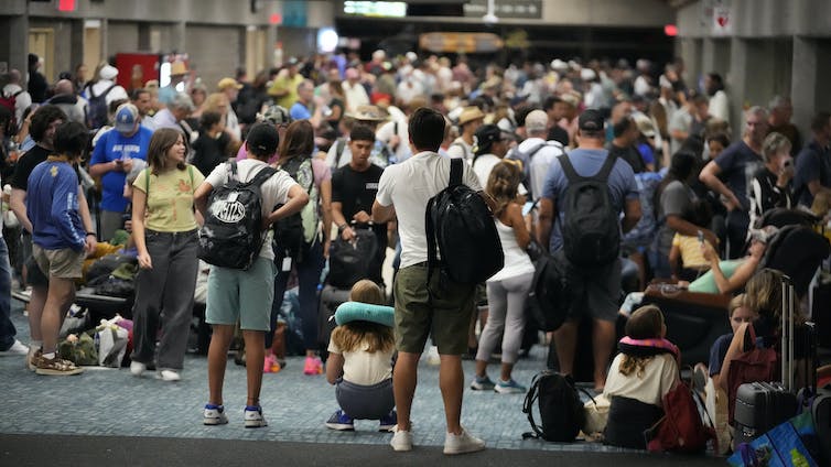 Large numbers of people, dressed for warm weather, mill about with luggage.