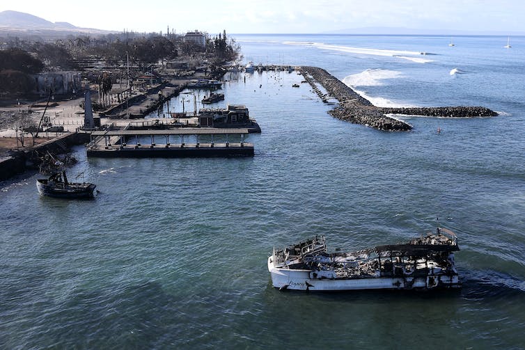 A tour boat and fishing boat are anchored near the coast, both severely burned, with the small harbor behind them.