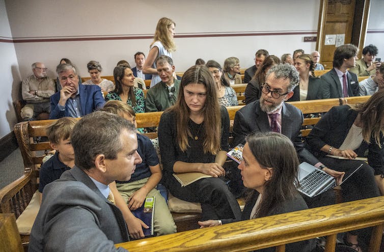 A young woman and two young boys listen as lawyers talk. Young people fill two rows of benches behind them in the small court room.