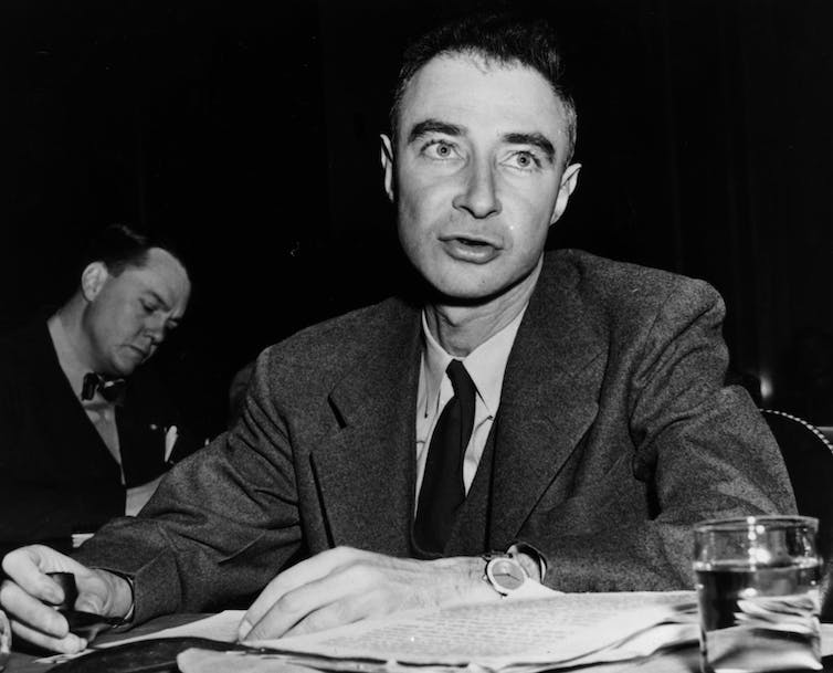 A man wearing a black jacket and tie, with his hands on a paper folder, speaking to an audience.