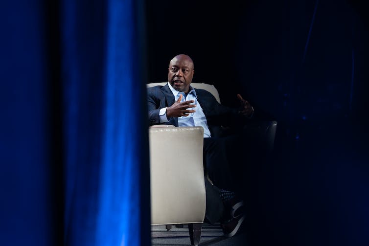 Tim Scott is seen, partially obscured by a blue curtain, sitting in a beige chair on a stage.