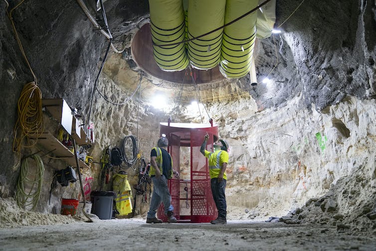 Workers in a rock cavern underground look up at a giant hole in the ceiling and pipe.