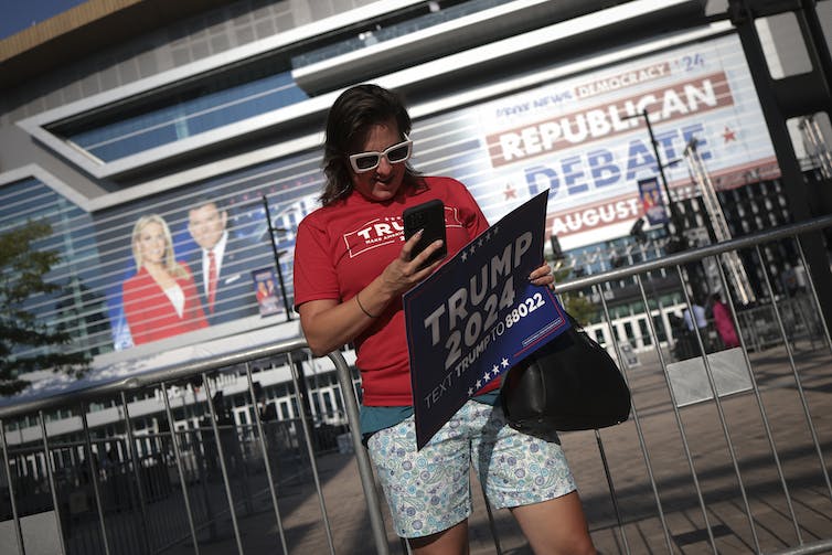 A dark-haired person in sunglasses and a red shirt holding a Trump sign in front of a large building.