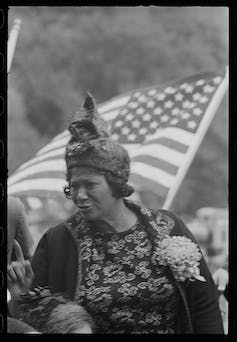 A Black woman wearing a black hat stands in front of an American flag.